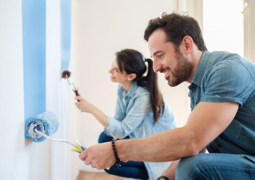 Two property managers smile as they paint the interior white wall of an investment property with blue paint.