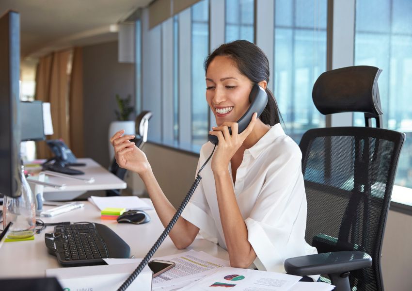 A remote landlord in a white blouse sits at a desk and speaks to their tenant on a landline telephone.