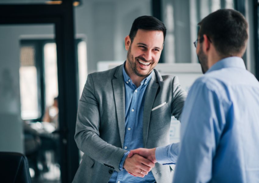 A smiling property manager in a blue button down and grey suit jacket shakes the hand of a landlord as they enter into partnership.