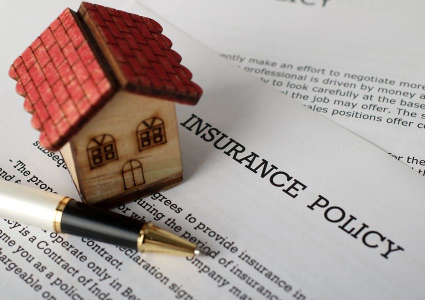 A small toy wooden house sits on top of a document that reads 'insurance policy'.