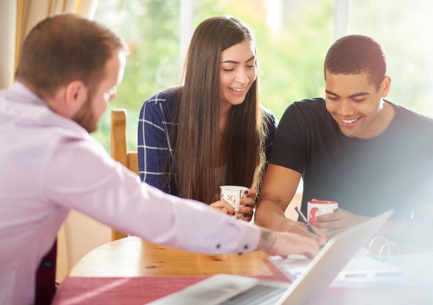 Two tenants sit at a table and sign a lease as directed by the landlord in front of them.