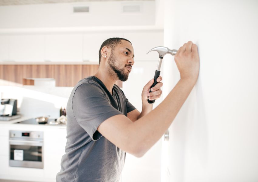 A self managing landord with a shaved head and beard uses a hammer against a wall during property maintenance.