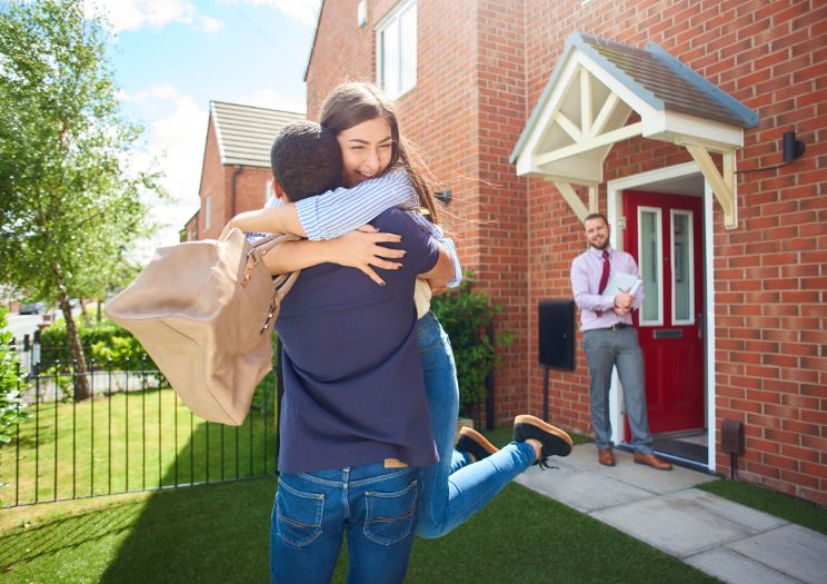 Two smiling and hugging tenants stand outside a rental property while their new landlord stands in the doorway.