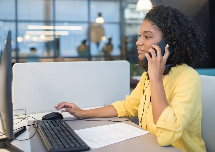 A property manager with medium length, dark curly hair and a yellow blouse sits at an office desk and speaks to a tenant on the phone.