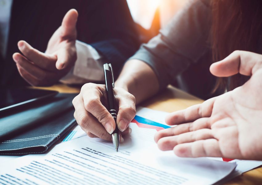 A close up of a desk with paperwork pictures one person with manicured nails filling out a form with two other hands gesture as if in conversation.