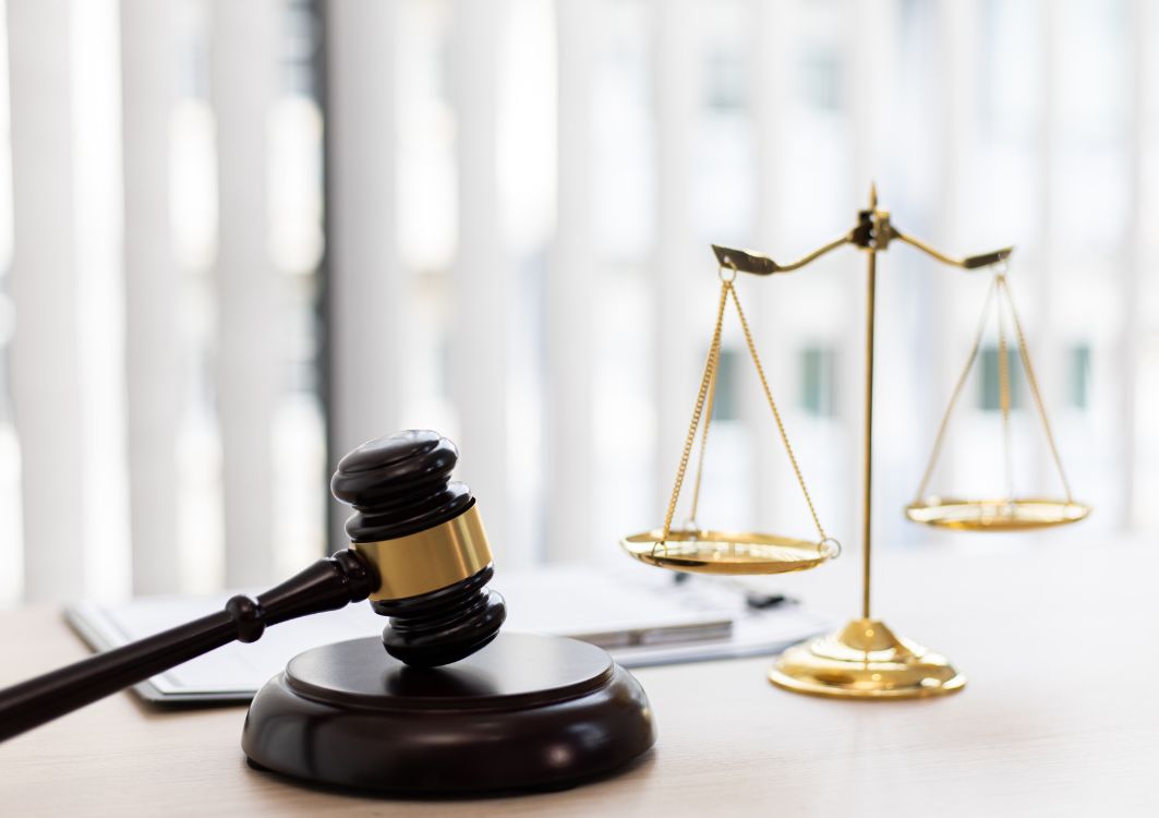 A brown and gold gavel tilts on a desk in front of a gold balancing scale.