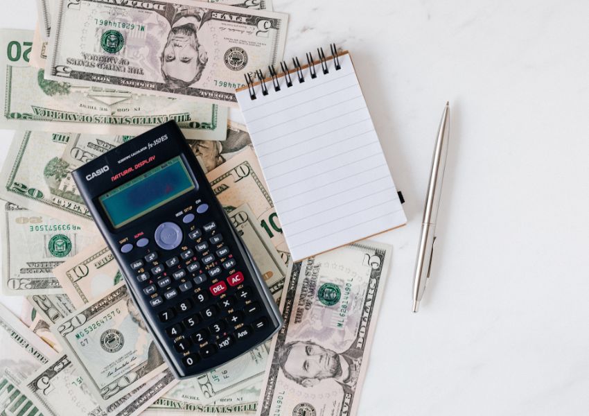 A black calculator, small notepad and silver pen sit on top of american bills on a white desk.